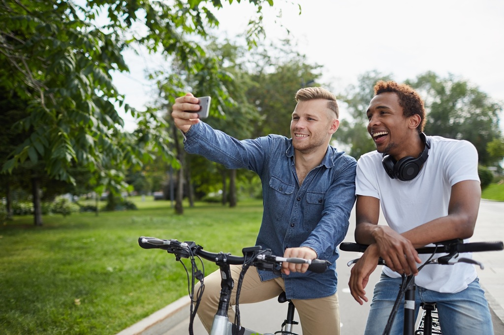 Two men taking a selfie on a phone as they sit on their bicycles outside on a park path